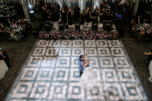 A bride and groom share their first dance on a striking geometric-patterned dance floor, illuminated by soft lighting. A live band performs in the background, framed by a vibrant floral arrangement, as wedding guests look on. The elegant setting, featuring luxurious décor and a romantic atmosphere, creates a breathtaking moment of celebration.