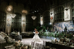 A bride and groom share their first dance in a grand, historic banquet hall adorned with gothic architecture, chandeliers, and intricate crests on the walls. The dimly lit space is illuminated by candlelight and warm ambient lighting, with a live band performing in front of a lush floral arrangement. Elegant tables set with fine dinnerware and a multi-tiered wedding cake complete the opulent setting.