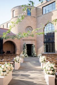 A beautiful outdoor wedding ceremony setup in a courtyard with warm brick architecture and large arched windows. White and blush floral arrangements line the aisle, leading to a doorway adorned with cascading flowers. Wooden cross-back chairs provide elegant seating, while greenery drapes overhead, adding a romantic touch to the sunlit space.