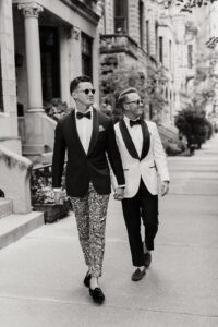 A stylish black and white photograph of two grooms walking hand in hand down a city sidewalk, dressed in elegant tuxedos with unique details—one in a classic white jacket and the other in a bold patterned pant ensemble. Both wear sunglasses and exude confidence and joy as they stroll past historic architecture, capturing a timeless and sophisticated wedding moment.