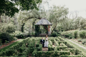 A bride and groom pose in a lush garden with a manicured hedge maze, surrounded by towering trees and greenery. The groom sits on a chair while the bride stands beside him, holding a bouquet. A charming brick and stone gazebo with a shingled roof serves as a picturesque backdrop, enhancing the romantic and timeless feel of the scene.