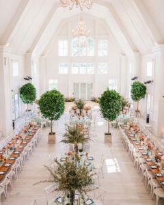 Elegant wedding reception setup in a bright, airy hall with high vaulted ceilings, large windows, and chandeliers. Long banquet tables adorned with floral arrangements, candles, and fine tableware line the room, complemented by lush green trees in planter boxes, creating a sophisticated and romantic atmosphere.