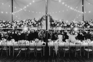 A black and white photograph of a lively wedding reception under a tent adorned with string lights. Guests are gathered, raising their hands in celebration beneath a long floral installation with lush white flowers and greenery. Elegant tables with candles, floral centerpieces, and Chiavari chairs add to the sophisticated ambiance of the event.